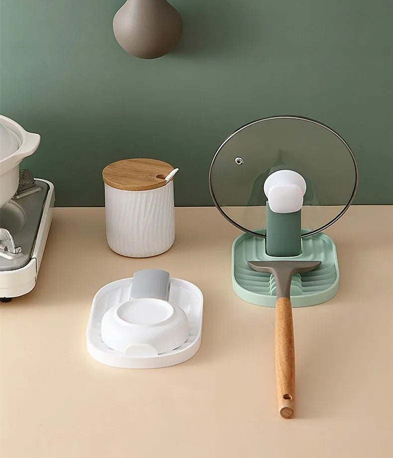 Silicone lid and spoon holder in use on a kitchen counter, holding a pot lid upright and a spatula on a grooved tray