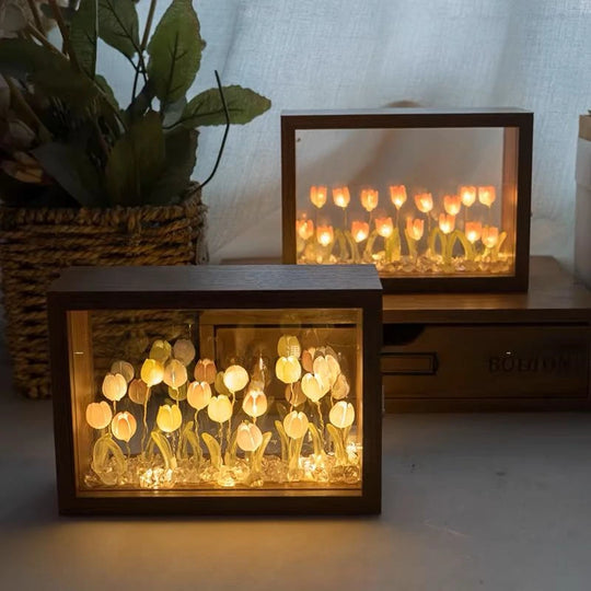 Two DIY wooden tulip light boxes with warm LED lighting, displaying soft yellow and peach-colored tulips on a table near a woven plant basket